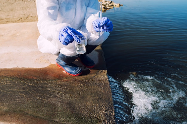 Ecologista irriconoscibile in piedi dove le acque reflue delle acque reflue incontrano il fiume e prelievo di campioni per determinare il livello di contaminazione e inquinamento