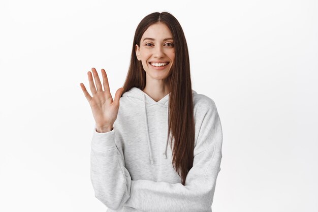 Ehi, come stai. Amichevole studentessa sorridente agitando la mano, salutando, benvenuto o salutandoti, facendo un gesto di addio, addio, in piedi su sfondo bianco