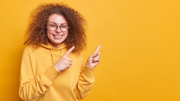 Ehi, guarda questo. Sorridente felice bella ragazza adolescente con capelli folti ricci invita a controllare la pubblicità mostra una bella offerta vestita in felpa isolata sul muro giallo dimostra il logo