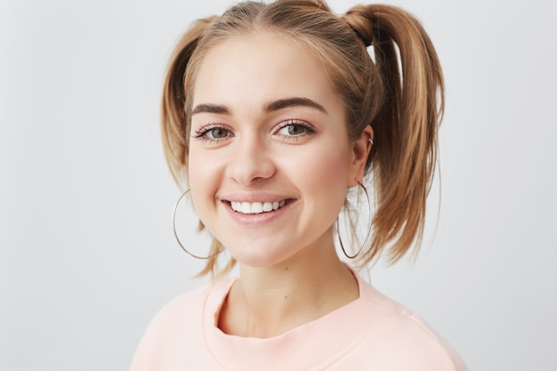 Elegante affascinante bella femmina caucasica con due code di cavallo, sorridendo felicemente, avendo gioia al chiuso. Bella donna positveyoung in maglione rosa che esprime emozioni e sentimenti positivi.