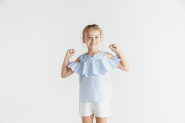 Elegante bambina sorridente in posa in abiti casual isolati su sfondo bianco studio. Modello femminile biondo caucasico. Emozioni umane, espressione facciale, infanzia. Vincere, festeggiare, sorridere.