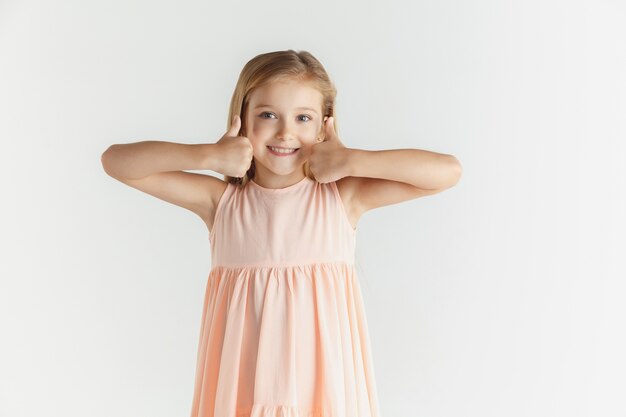 Elegante bambina sorridente in posa in abito isolato sul muro bianco. Modello femminile biondo caucasico. Emozioni umane, espressione facciale, infanzia. Sembra calmo, mostrando il segno di simpatia.