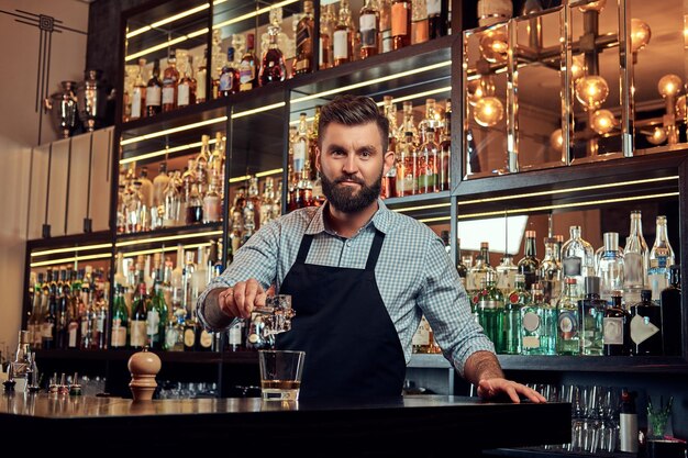 Elegante barista brutale in camicia e grembiule fa un cocktail sullo sfondo del bancone del bar.