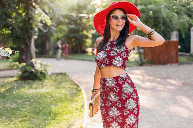 Elegante bella donna che cammina nel parco in abito tropicale. signora nella tendenza della moda estiva street style. indossa una borsa di paglia, cappello rosso, occhiali da sole, accessori. ragazza sorridente di buon umore in vacanza.