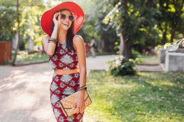 Elegante bella donna che cammina nel parco in abito tropicale. signora nella tendenza della moda estiva street style. indossa una borsa di paglia, cappello rosso, occhiali da sole, accessori. ragazza sorridente di buon umore in vacanza.