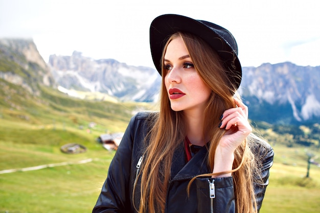 Elegante bella donna con i capelli lunghi, indossa un cappello alla moda e giacca di pelle, in posa alle Alpi Dolomiti italiane