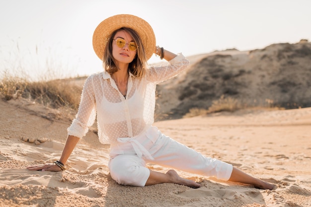 Elegante bella donna nella sabbia del deserto in abito bianco che indossa il cappello di paglia sul tramonto