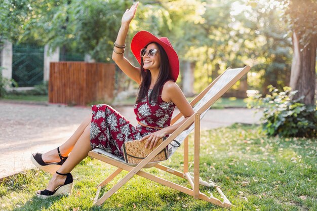Elegante bella donna seduta sulla sedia a sdraio in abito stile tropicale, agitando la mano, tendenza moda estiva, borsa di paglia, cappello rosso, occhiali da sole, accessori, sorridente, umore felice, vacanze