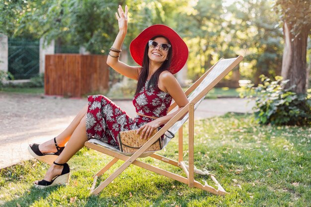 Elegante bella donna seduta sulla sedia a sdraio in abito stile tropicale, agitando la mano, tendenza moda estiva, borsa di paglia, cappello rosso, occhiali da sole, accessori, sorridente, umore felice, vacanze