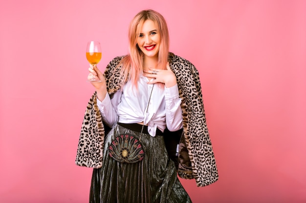 Elegante bella giovane donna positiva che si diverte, indossa un abito da cocktail scintillante da sera e un cappotto alla moda con stampa leopardata di pelliccia, sfondo rosa, godendosi la festa delle vacanze invernali.