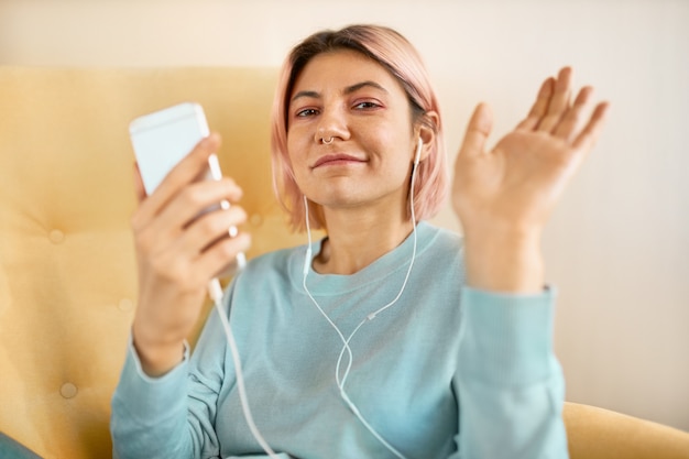 Elegante bella ragazza in auricolari seduto in poltrona agitando la mano alla telecamera, dicendo ciao