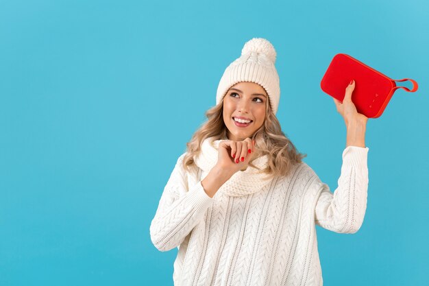 Elegante bionda sorridente bella giovane donna che tiene altoparlante wireless ascoltando musica ballare felice indossando maglione bianco e cappello lavorato a maglia moda stile invernale in posa isolato sulla parete blu
