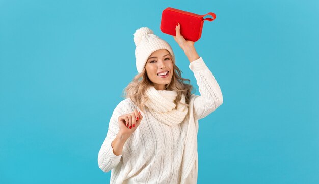 Elegante bionda sorridente bella giovane donna che tiene altoparlante wireless ascoltando musica che indossa un maglione bianco e cappello lavorato a maglia in posa sul blu