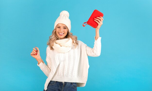 Elegante bionda sorridente bella giovane donna che tiene altoparlante wireless ascoltando musica che indossa un maglione bianco e cappello lavorato a maglia in posa sul blu