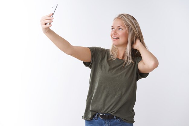 Elegante blogger di lifestyle femminile che prende selfie sullo smartphone controllando il taglio di capelli allunga la mano con il telefono cellulare scatta una buona ripresa parlando videochiamata su sfondo bianco, sorridendo allo schermo del gadget