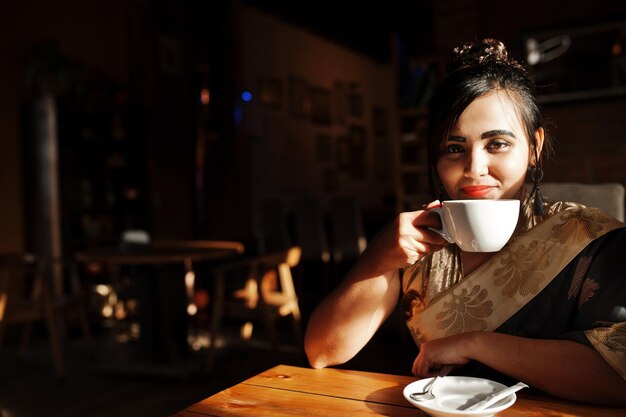 Elegante bruna ragazza indiana del sud asiatico in saree poste caffè al coperto e bere il tè