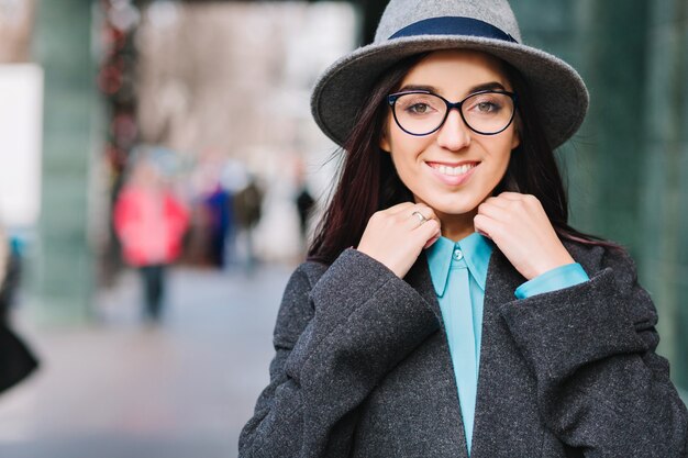 Elegante città ritratto giovane donna graziosa con cappello grigio, occhiali neri che camminano sulla strada nel centro. Cappotto di lusso, modello alla moda, emozioni allegre, sorridente.
