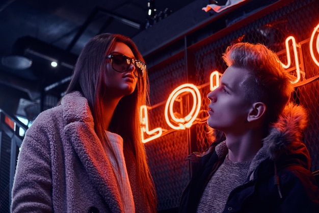 Elegante coppia che indossa abiti caldi in piedi nella caffetteria con interni industriali, un'insegna retroilluminata sullo sfondo