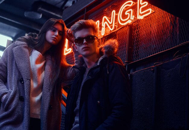 Elegante coppia che indossa abiti caldi in piedi sulle scale per la discoteca sotterranea, un cartello retroilluminato sullo sfondo