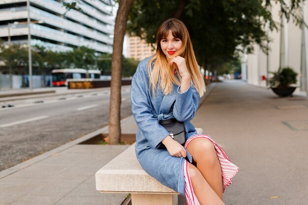 Elegante donna abbastanza felice in cappotto blu che si siede sulla panchina in strada