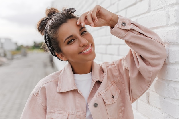 Elegante donna adolescente dai capelli scuri in giacca di jeans alla moda si è appoggiata al muro di mattoni bianchi