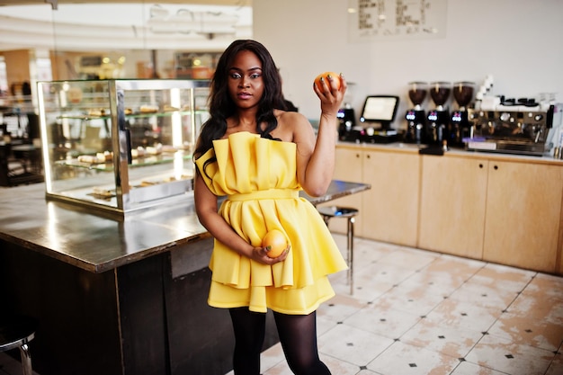 Elegante donna afroamericana in abito giallo posata al caffè con due pompelmi sulle mani