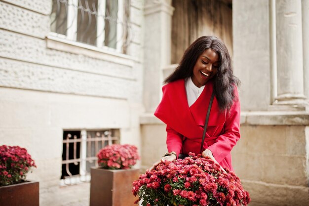 Elegante donna afroamericana in cappotto rosso posata contro i fiori all'aperto