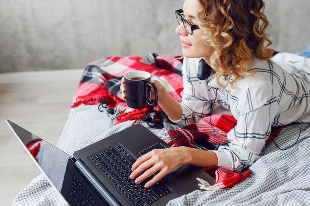 Elegante donna allegra intelligente in occhiali utilizzando il computer portatile, sdraiato sul letto accogliente, bere caffè.