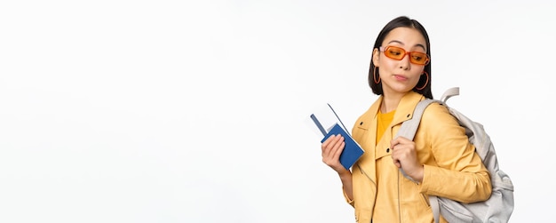 Elegante donna asiatica in occhiali da sole per andare all'estero con zaino turistico in possesso di passaporto e biglietti d'imbarco aereo in piedi su sfondo bianco