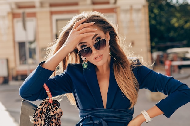 Elegante donna attraente che indossa abito elegante blu e occhiali da sole camminando nella borsa della holding della strada