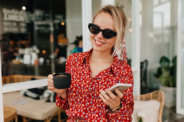 Elegante donna bionda che indossa abiti estivi luminosi utilizzando smartphone e bere caffè all'esterno