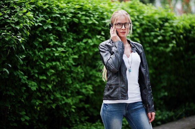 Elegante donna bionda indossa jeans, occhiali e giacca di pelle con telefono a portata di mano contro i cespugli in strada Ritratto di modello urbano di moda