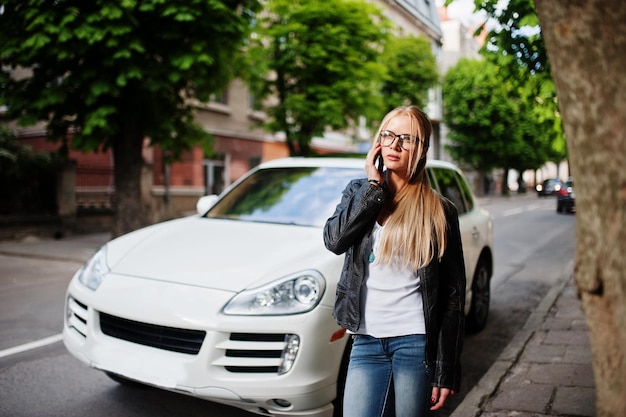 Elegante donna bionda indossare jeans, occhiali e giacca di pelle con telefono cellulare contro auto di lusso Ritratto di modello urbano di moda