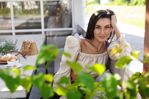 Elegante donna bruna sensuale magra sul divano alla macchina fotografica del sorriso del coffeeshop Donna attraente che cena fuori al caffè che beve caffè all'aperto nel ristorante Signora sexy che ha appuntamento romantico o pranzo