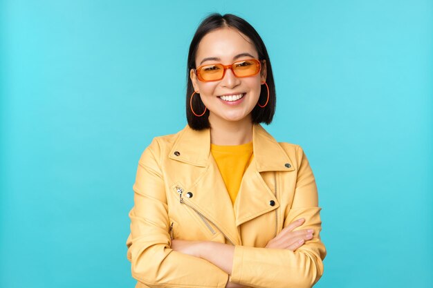 Elegante donna coreana in occhiali da sole e giacca gialla incrocia le braccia sorridendo e guardando fiduciosa in telecamera in piedi su sfondo blu