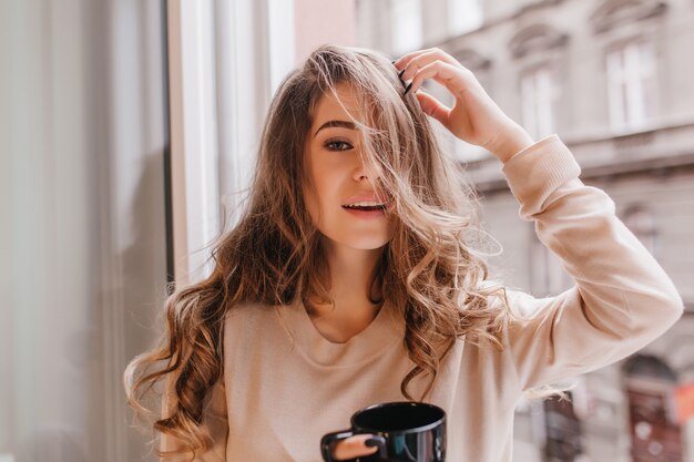 Elegante donna dai capelli lunghi in posa al mattino, bevendo caffè accanto alla finestra