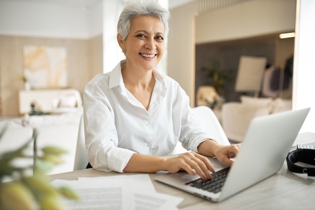 Elegante donna di affari matura con taglio di capelli corto seduto davanti al computer portatile, guardando lo schermo con la bocca aperta come se dicesse qualcosa