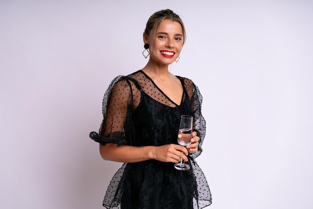 Elegante donna elegante che indossa un abito da sera nero che tiene in mano un bicchiere con un drink, pronto per la celebrazione, sfondo bianco.