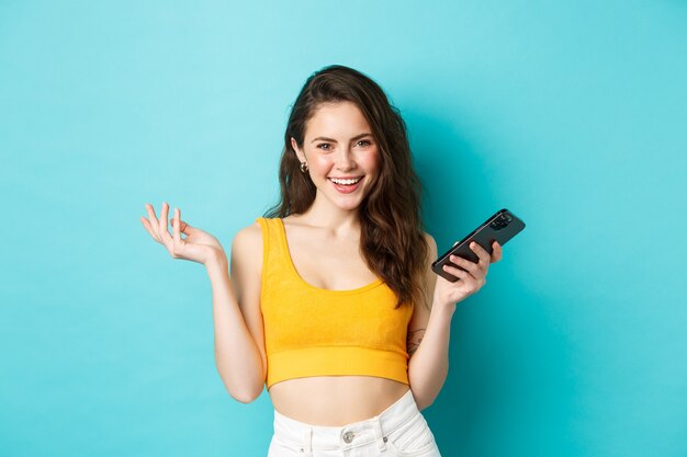 Elegante donna felice che tiene smartphone, ride e sorride sicura di sé, chiacchiera al telefono cellulare, in piedi su sfondo blu.
