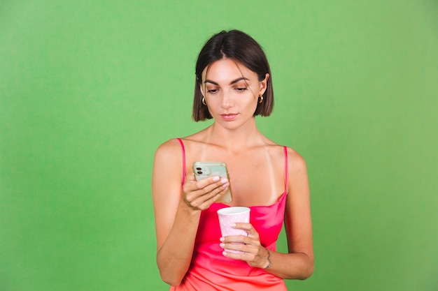 Elegante donna in abito di seta rosa isolata su verde felice con un sorriso guarda sullo schermo del telefono cellulare, leggi le notizie sui messaggi