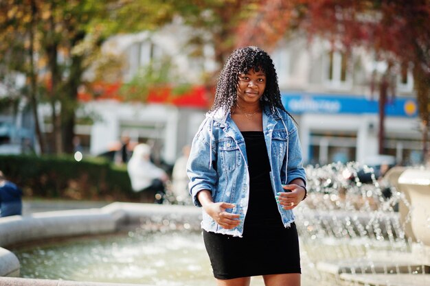 Elegante donna riccia afro francese alla moda posata al giorno d'autunno in giacca di jeans Modello femminile africano nero contro fontana