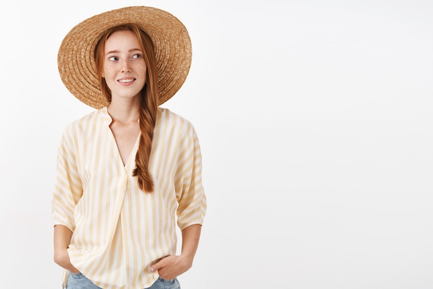 Elegante donna rossa che va in spiaggia indossando cappello di paglia per non abbronzarsi girando a destra con felice espressione spensierata tenendo le mani in tasca godendo di una calda e soleggiata giornata estiva