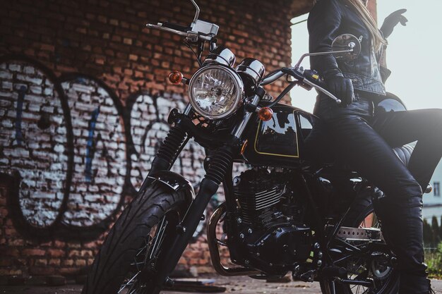 Elegante donna sexy in abbigliamento da motociclista sta posando per il fotografo accanto alla sua bici e al muro dei graffiti.