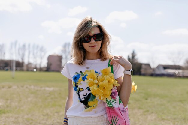 Elegante donna ucraina con un taglio di capelli corto e leggero è vestita maglietta bianca con stampa blu e gialla tiene in mano una borsa con fiori gialli su sfondo naturale