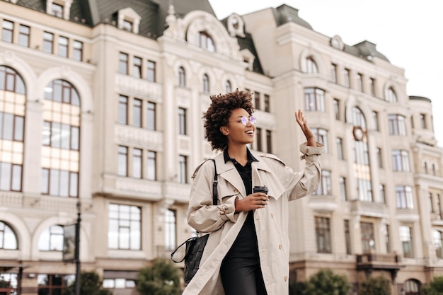 Elegante e affascinante bruna signora dalla carnagione scura in occhiali da vista trench beige e vestito nero sorride onde la mano in segno di saluto e tiene una tazza di caffè all'aperto