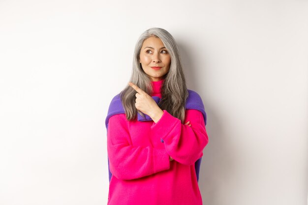 Elegante e moderna donna anziana asiatica che punta all'angolo in alto a sinistra, guardando il logo con un sorriso compiaciuto, in piedi con un maglione rosa su sfondo bianco