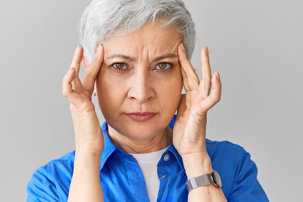 Elegante femmina caucasica di mezza età in camicia blu che soffre di emicrania. Immagine ravvicinata di donna matura stressata che stringe le tempie a causa di un terribile mal di testa, massaggiando i punti dolorosi