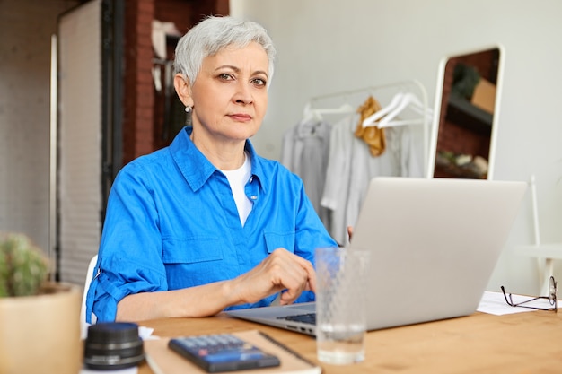 Elegante fotografa dai capelli grigi sulla sessantina, seduta in ufficio davanti al computer portatile aperto, caricando le immagini. Donna matura che naviga in Internet utilizzando un gadget elettronico generico