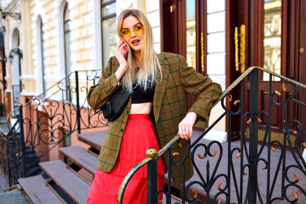 Elegante giovane donna bionda in posa vicino al negozio di lusso e parlando dal suo smartphone, vestito moderno alla moda, giacca oversize e marsupio.