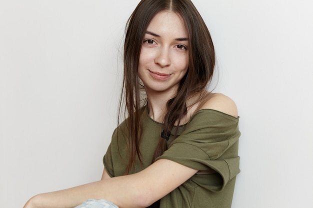 Elegante giovane donna caucasica con acconciatura disordinata che indossa top verde militare oversize, guardando con un sorriso felice e gioioso, trascorrendo il suo giorno libero al chiuso. Concetto di persone e stile di vita